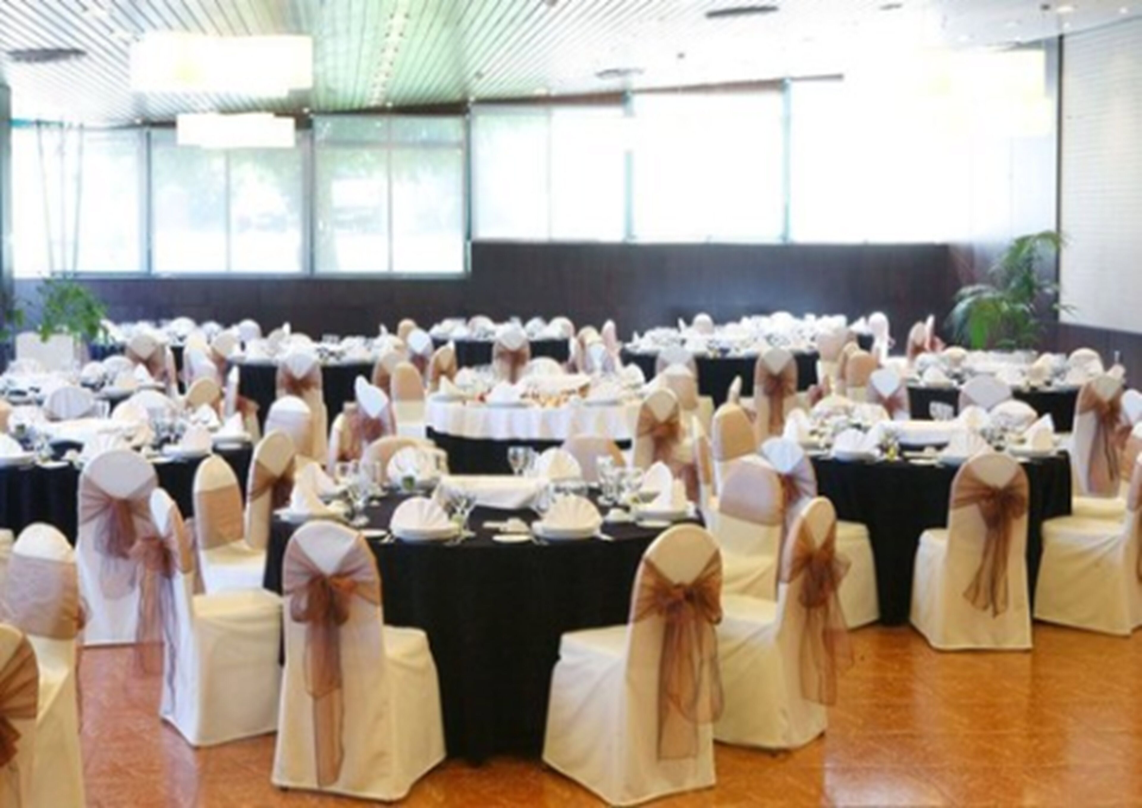 Un salón de banquetes con mesas redondas dispuestas para un evento formal, con manteles blancos y sillas con lazos marrones.