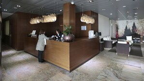 A reception desk with a wooden finish, a large potted plant, and a wall with a cityscape mural. - Hotel Spa Congreso (Teo)