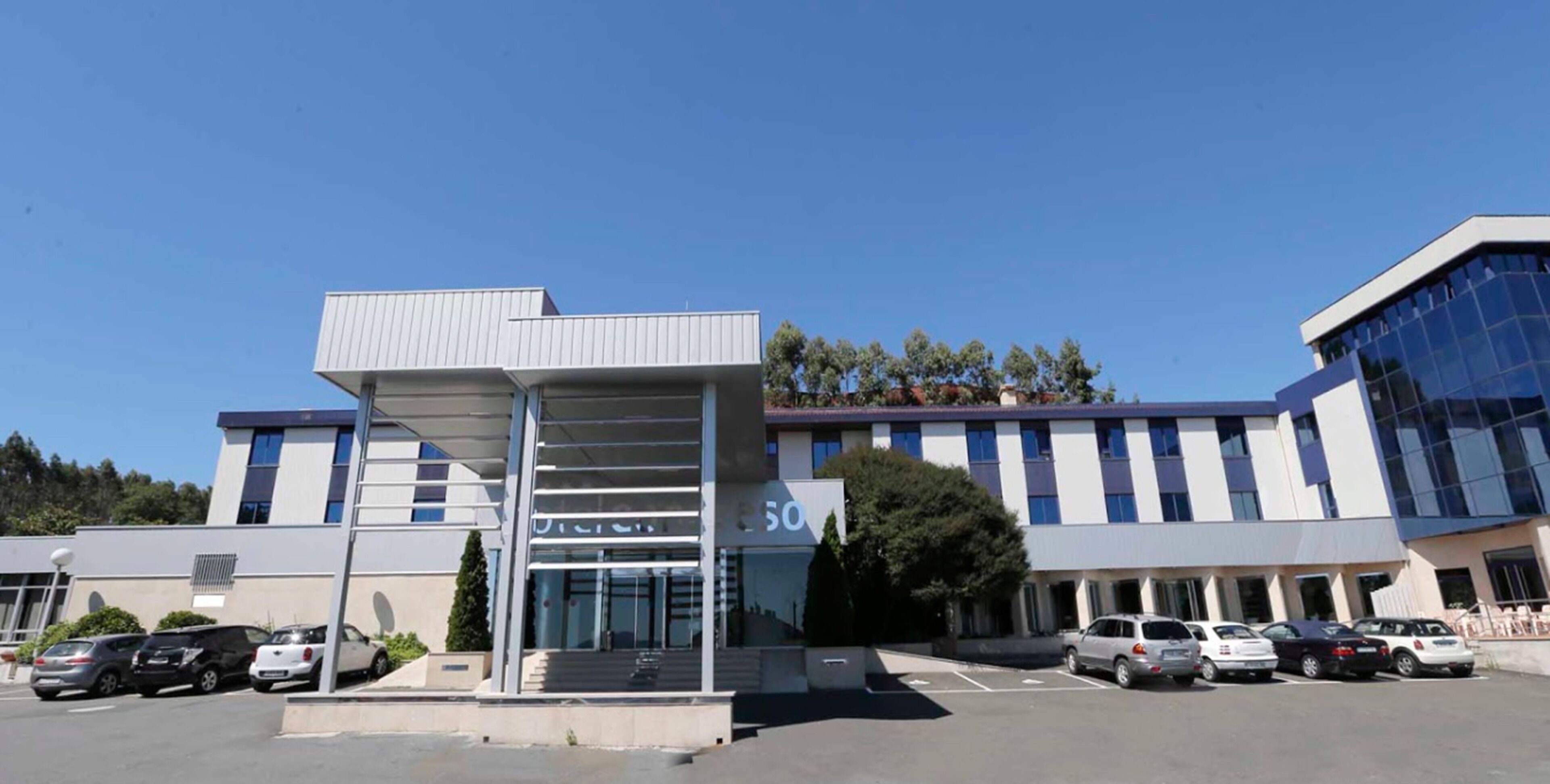 Un edificio moderno con una entrada destacada, un estacionamiento con varios autos y un cielo azul despejado.