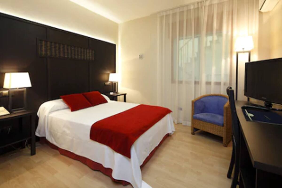 Une chambre d’hôtel avec un grand lit, deux tables de chevet avec des lampes, une télévision, un fauteuil en osier et une fenêtre avec des rideaux légers.