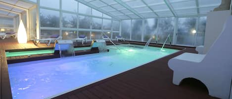 An indoor swimming pool with clear glass walls, lounge chairs, and a statue.