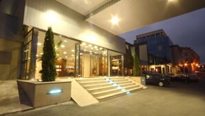 A hotel entrance with a glass facade, outdoor seating area, and a staircase leading to the entrance. - Hotel Spa Congreso (Teo)