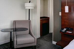 Quarto de hotel com uma poltrona cinza, uma mesa redonda pequena, um abajur, um telefone e um painel de madeira na parede.