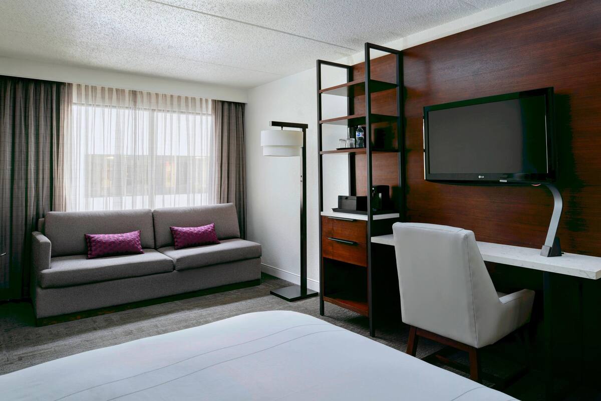 A hotel room with a bed, a sofa, a desk, a chair, a TV, and a window with curtains.