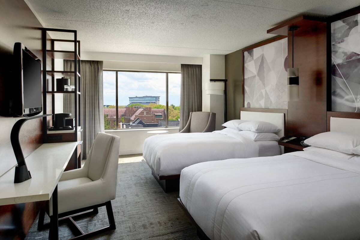 A hotel room with two beds, a desk, a chair, a TV, and a view of a building outside.