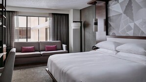 A modern hotel room with a large bed, a sofa with cushions, a desk, and a large window with curtains. - Marriott East Lansing at University Place (East Lansing)