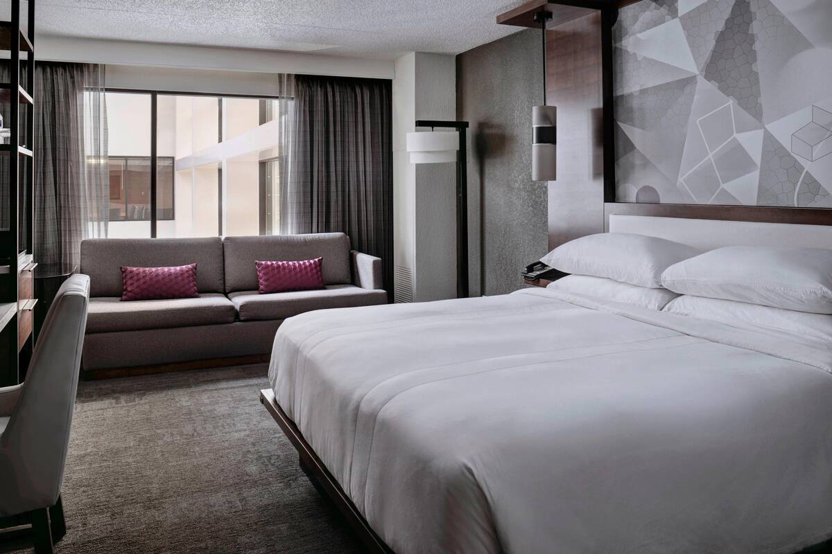 A modern hotel room with a large bed, a sofa with cushions, a desk, and a large window with curtains.