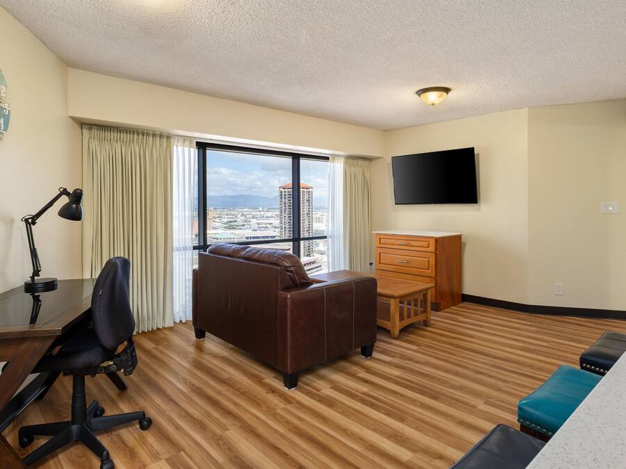 A hotel room with a large window, a desk with a chair, a sofa, a dresser, and a television on the wall.