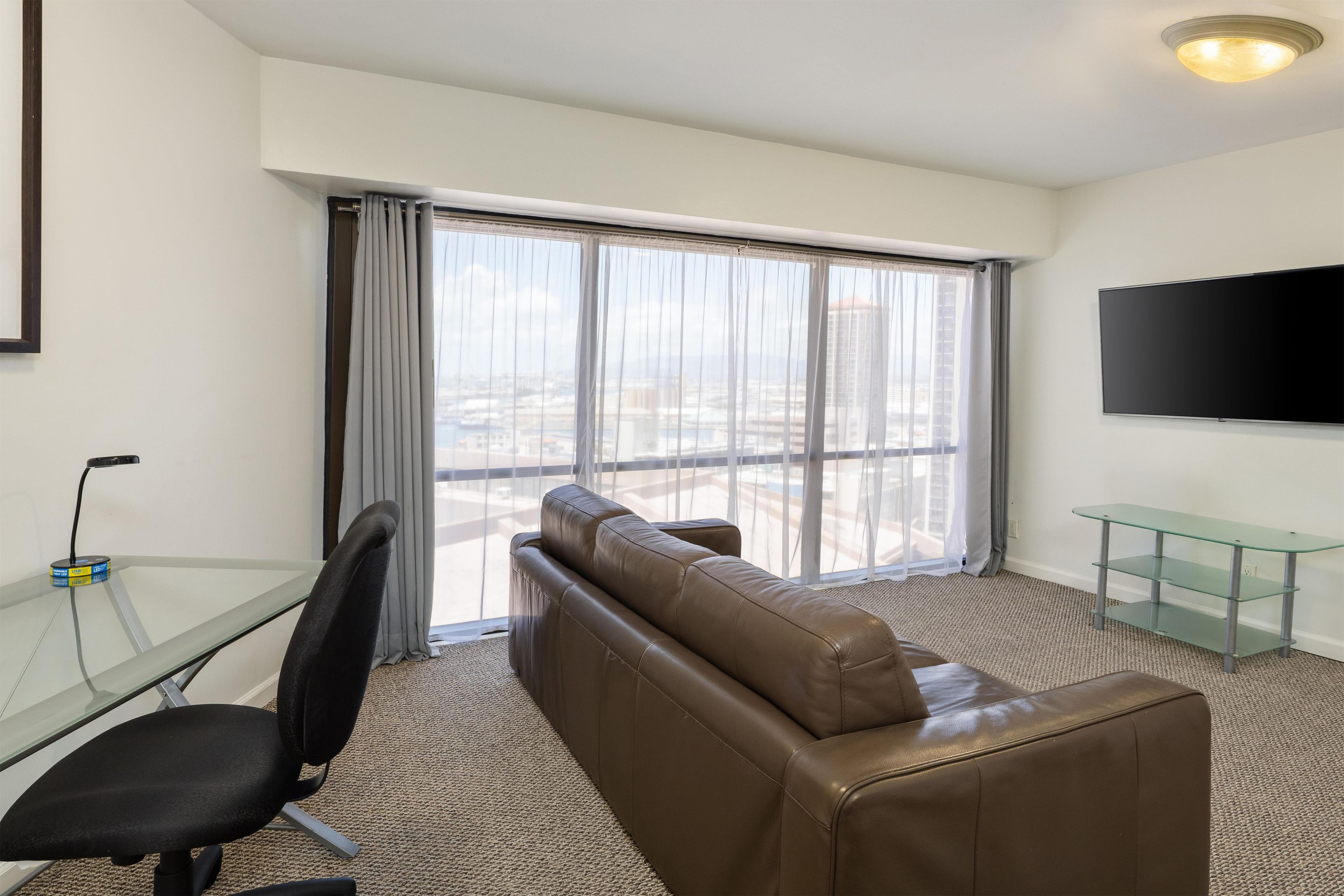 A hotel room with a brown leather sofa, a glass desk with a chair, a flat-screen TV mounted on the wall, and a large window with curtains.