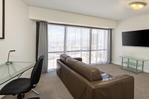 A hotel room with a brown leather sofa, a glass desk with a chair, a flat-screen TV mounted on the wall, and a large window with curtains. - Aston at the Executive Centre Hotel (Honolulu)