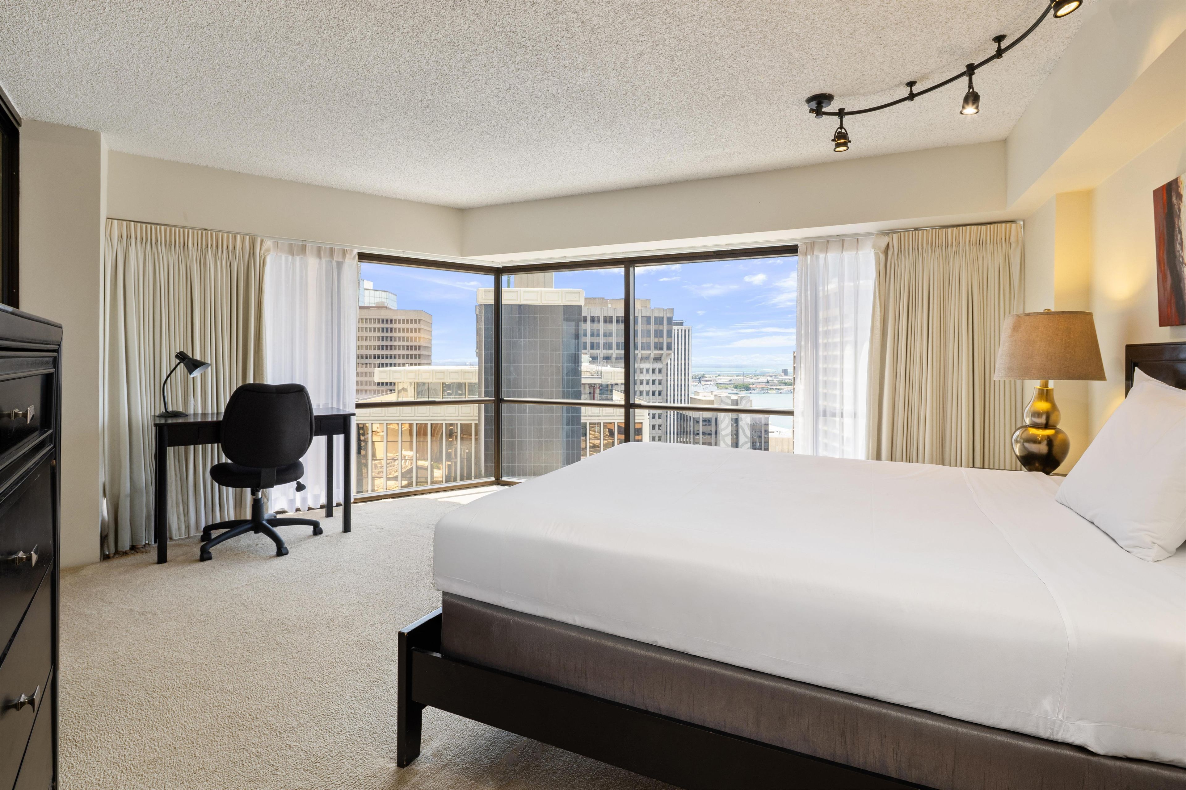 A hotel room with a large bed, a desk, a chair, and a view of the cityscape through the window.
