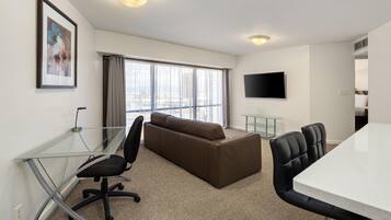 A hotel room with a glass desk, a brown sofa, a TV mounted on the wall, and a large window with curtains.