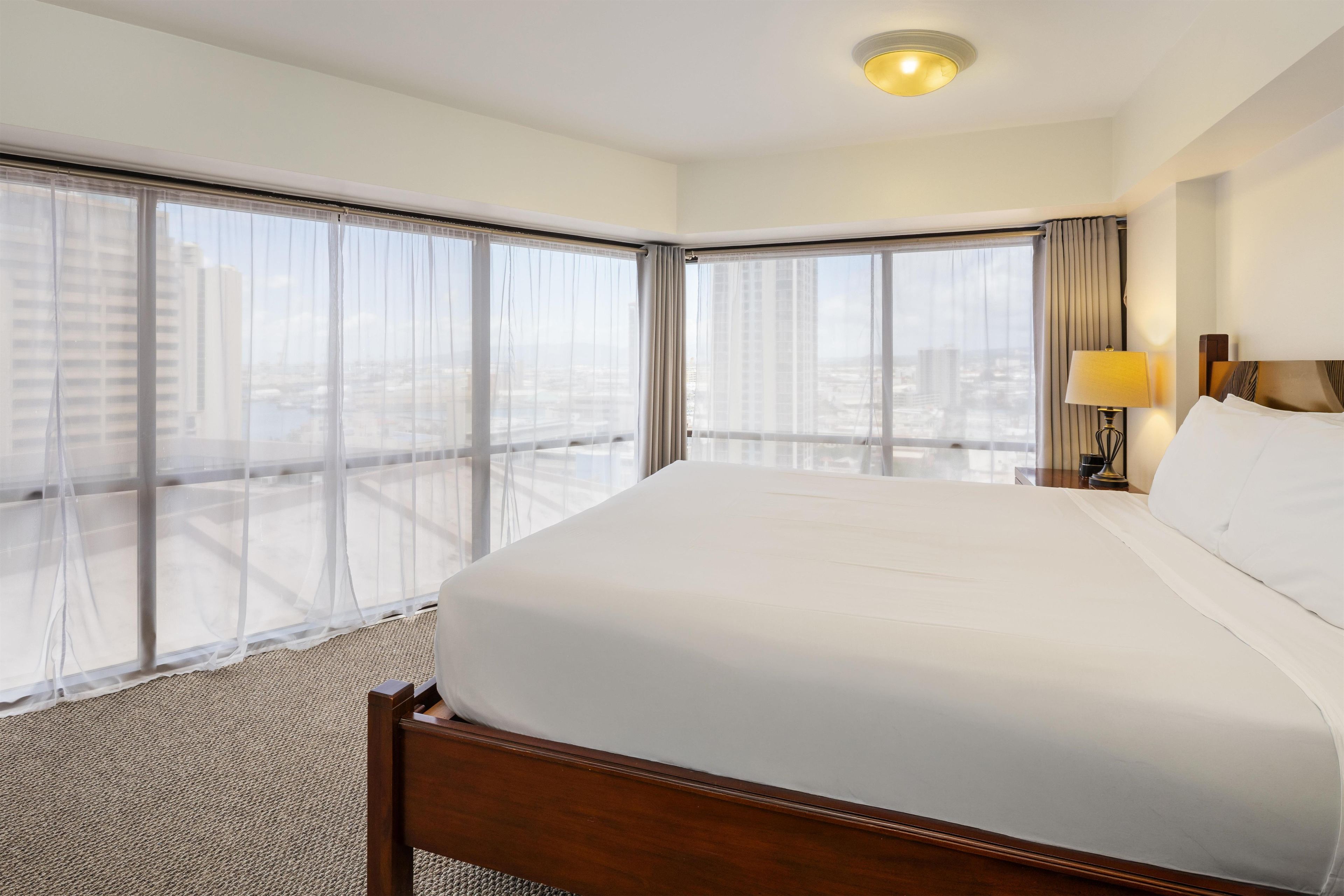 Habitación de hotel con una cama grande, una mesita de noche, una lámpara y vistas a la ciudad a través de ventanales de gran altura.