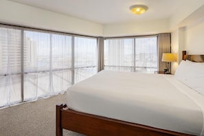 A hotel room with a large bed, a nightstand, a lamp, and a view of the city through floor-to-ceiling windows. - Aston at the Executive Centre Hotel (Honolulu)
