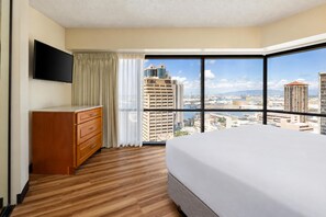 A hotel room with a large bed, a wooden dresser, a flat-screen TV, and a city view through large windows. - Aston at the Executive Centre Hotel (Honolulu)