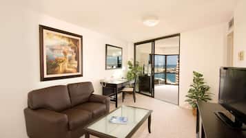 A living room with a brown leather sofa, a glass coffee table, a desk with a chair, a TV, and a large window with a view of the sea.