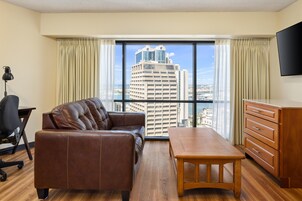 A hotel room with a leather sofa, a wooden table, a desk with a lamp, a TV, and a view of a cityscape through large windows.