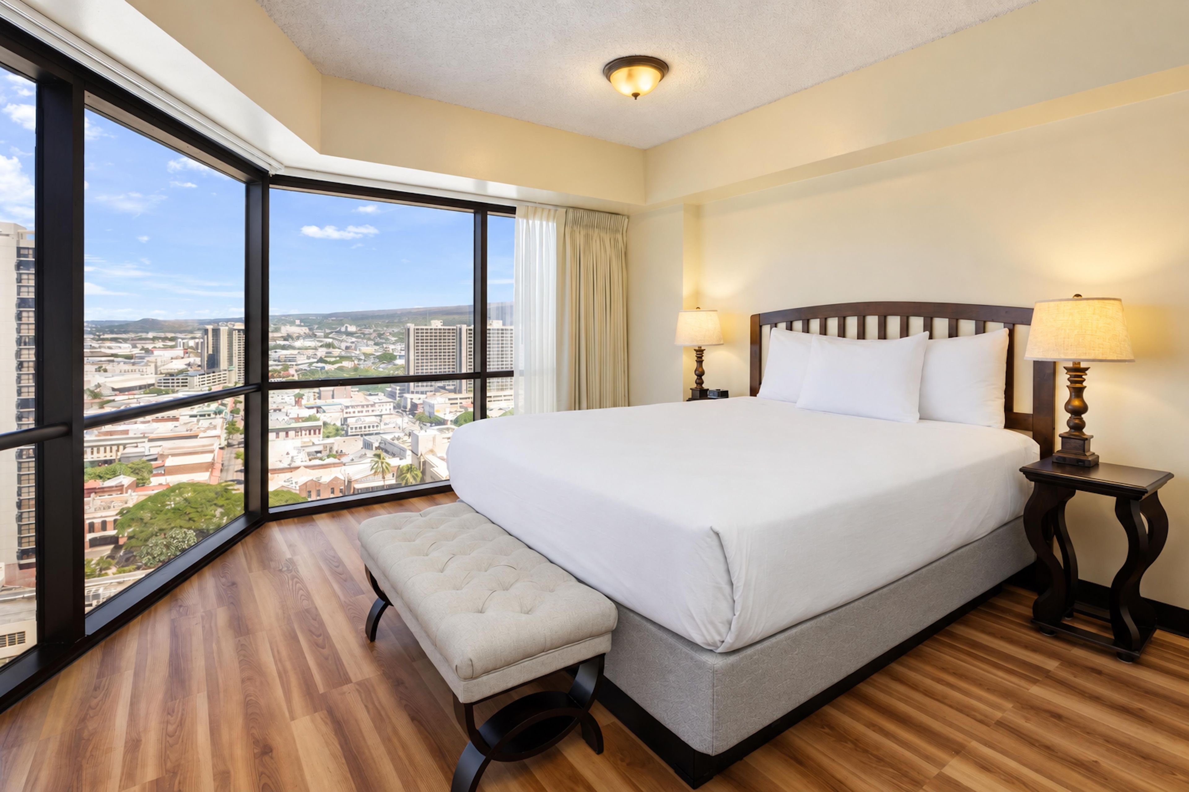 A hotel room with a large bed, a bench, a nightstand, and a view of the city.