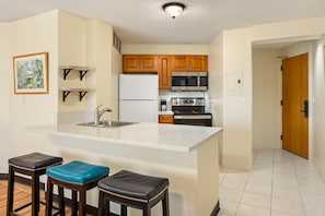 A kitchen with a central island, wooden cabinets, a microwave, a refrigerator, and a coffee maker. There are two stools with blue cushions. - Aston at the Executive Centre Hotel (Honolulu)