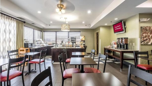 A modern dining area with a kitchen in the background, a flat-screen TV displaying "Free Wi-Fi All Rooms", and a ceiling fan. - Sonesta Essential Ames (Ames)