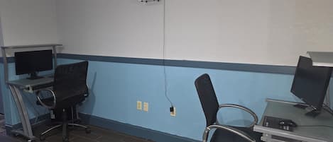 A room with two desks, each with a computer monitor and keyboard, and two black office chairs. There is a whiteboard on the wall and a power outlet near the center.