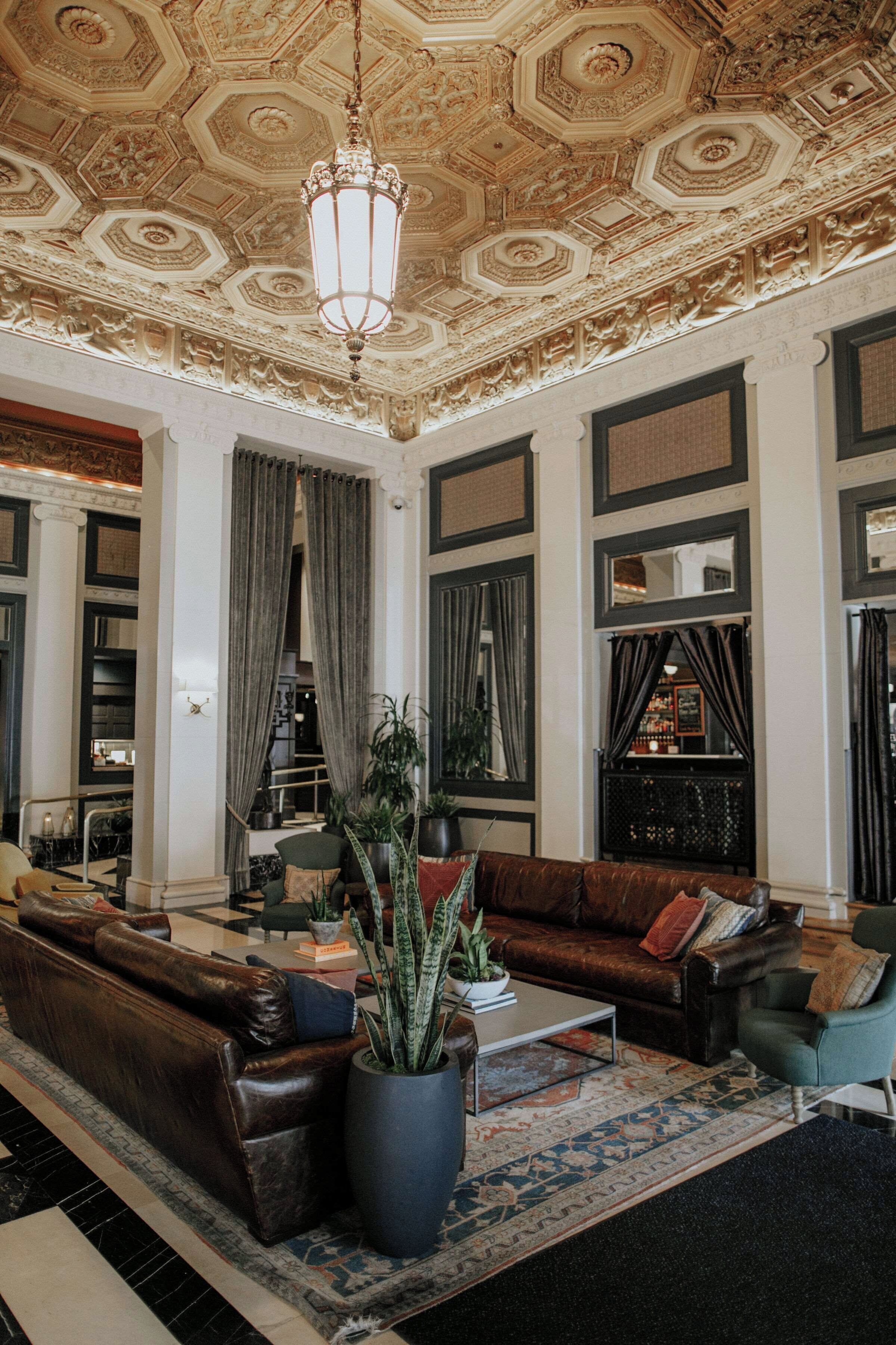 A spacious lobby with a high ornate ceiling, leather sofas, a coffee table, and a large potted plant.