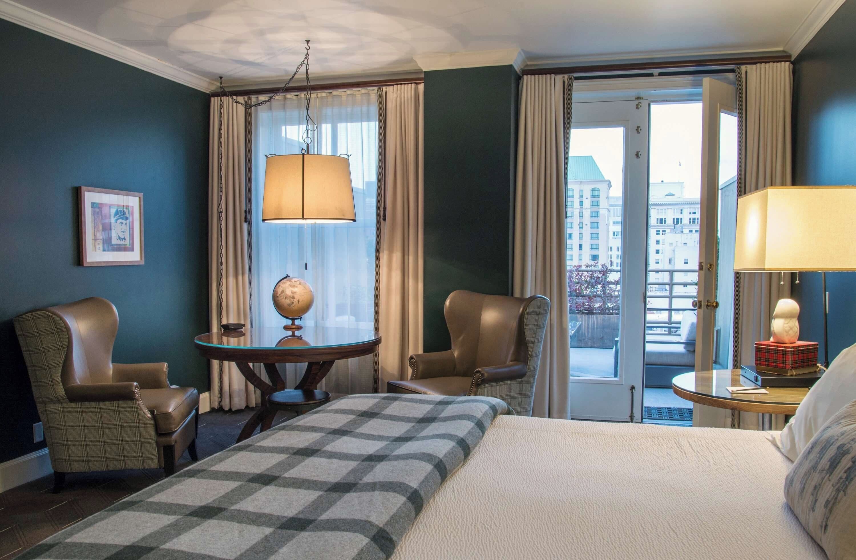 Hotelkamer met een bed, twee stoelen, een klein tafeltje met een wereldbol, een lamp en uitzicht op de stad door de deur.