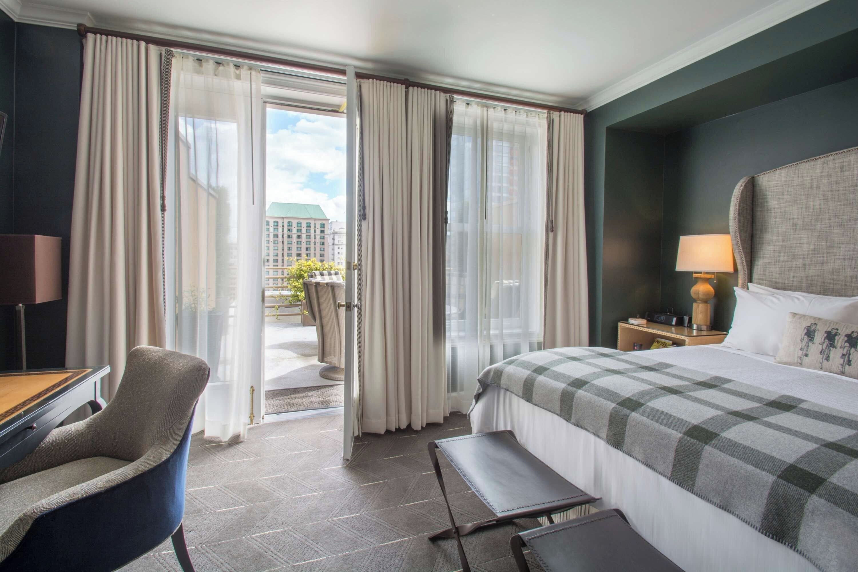 A hotel room with a bed, a desk, a chair, a nightstand with a lamp, and a view of the city through the window.