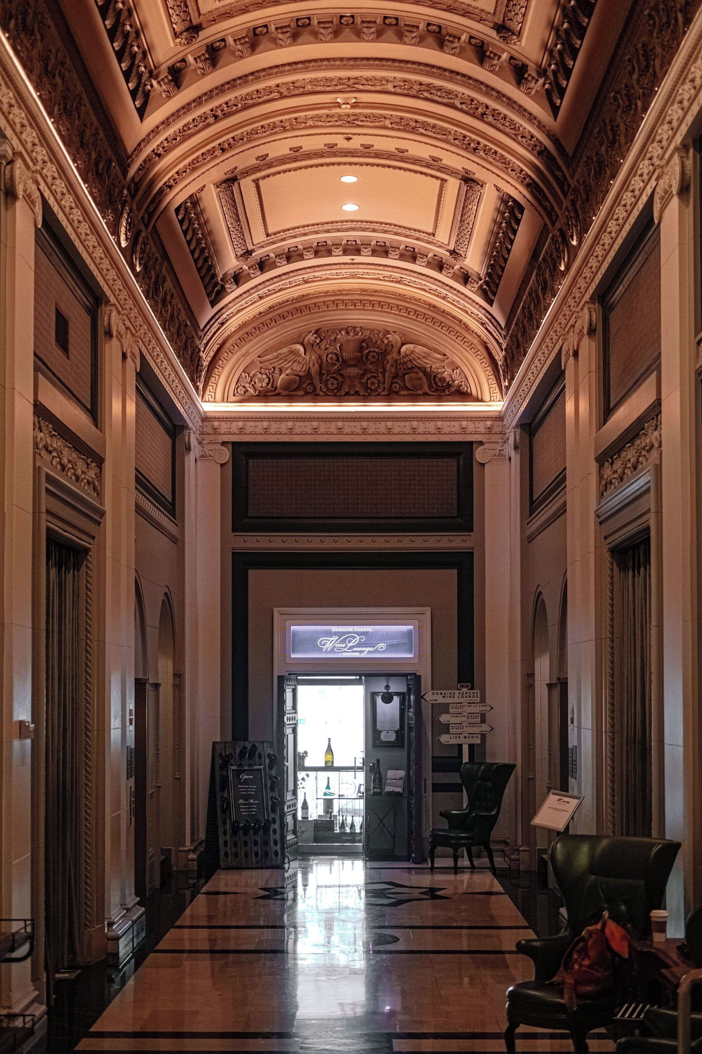 A grand lobby with ornate ceiling and columns, a wine lounge sign, and a seating area with chairs.