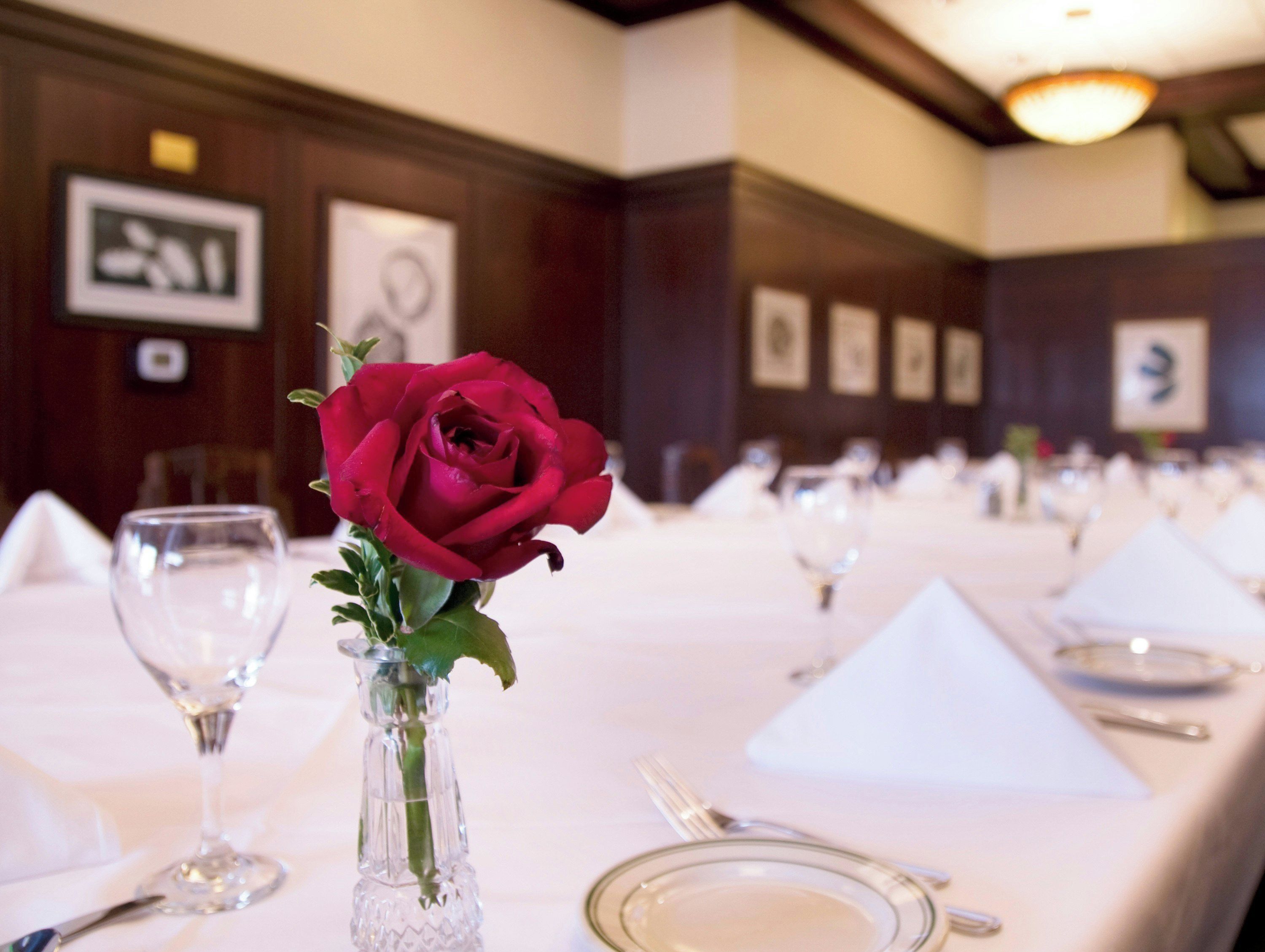A dining table set with a clear glass vase containing a single red rose, surrounded by white tablecloths, plates, and cutlery.