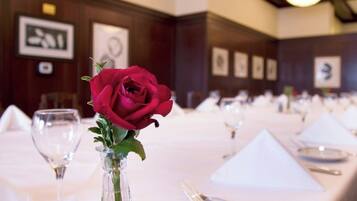 A dining table set with a clear glass vase containing a single red rose, surrounded by white tablecloths, plates, and cutlery.
