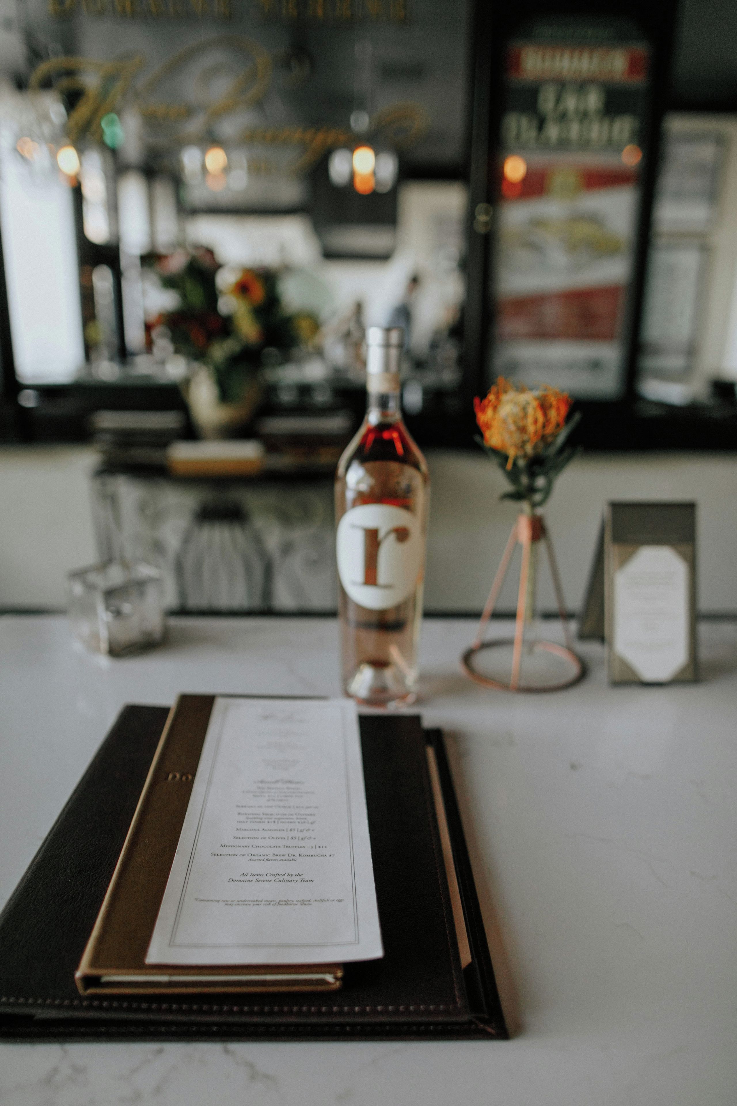 A menu on a black tray with a bottle of liquor and a vase of flowers on a table.