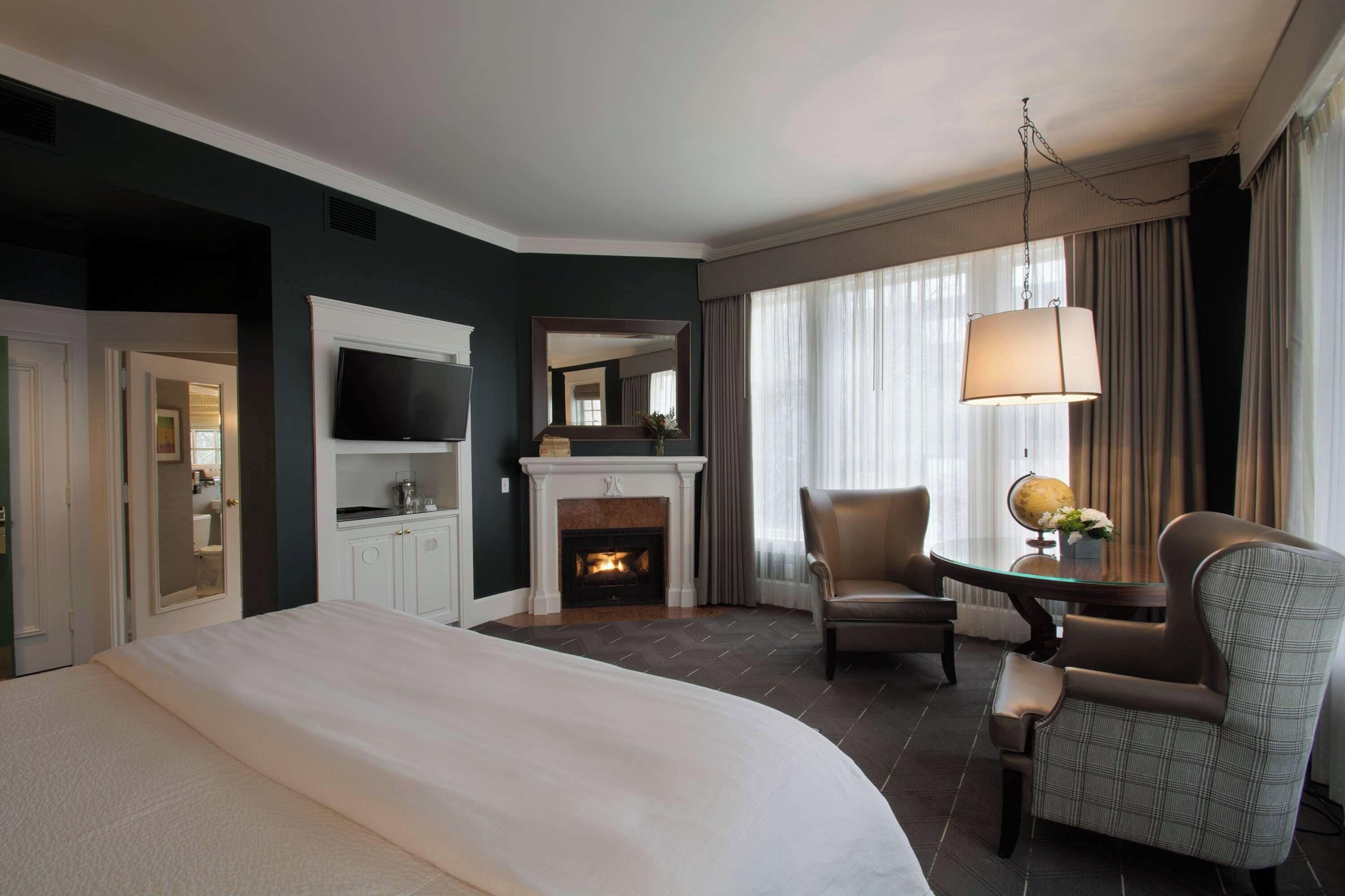 A hotel room with a bed, a television, a fireplace, a desk with a lamp, and a chair.