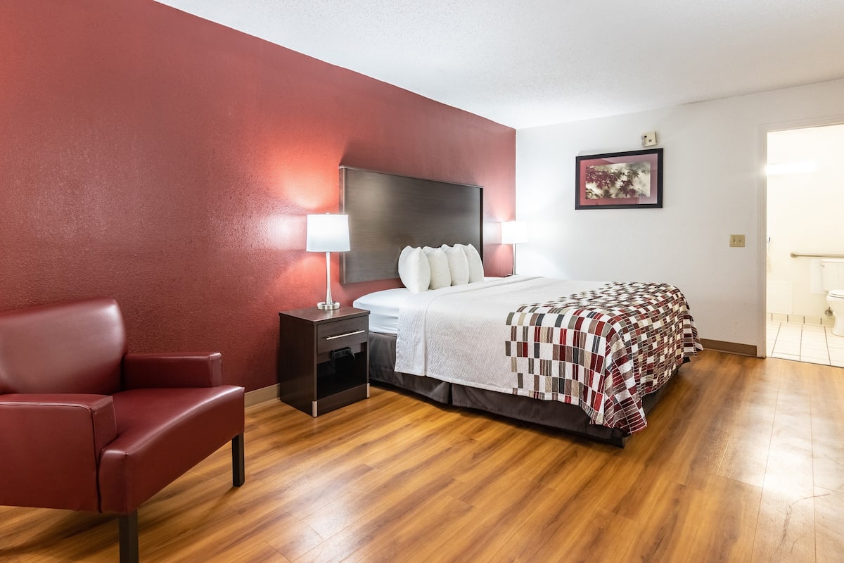 Una habitación de hotel con una cama, una mesita de noche, una silla roja y suelo de madera.