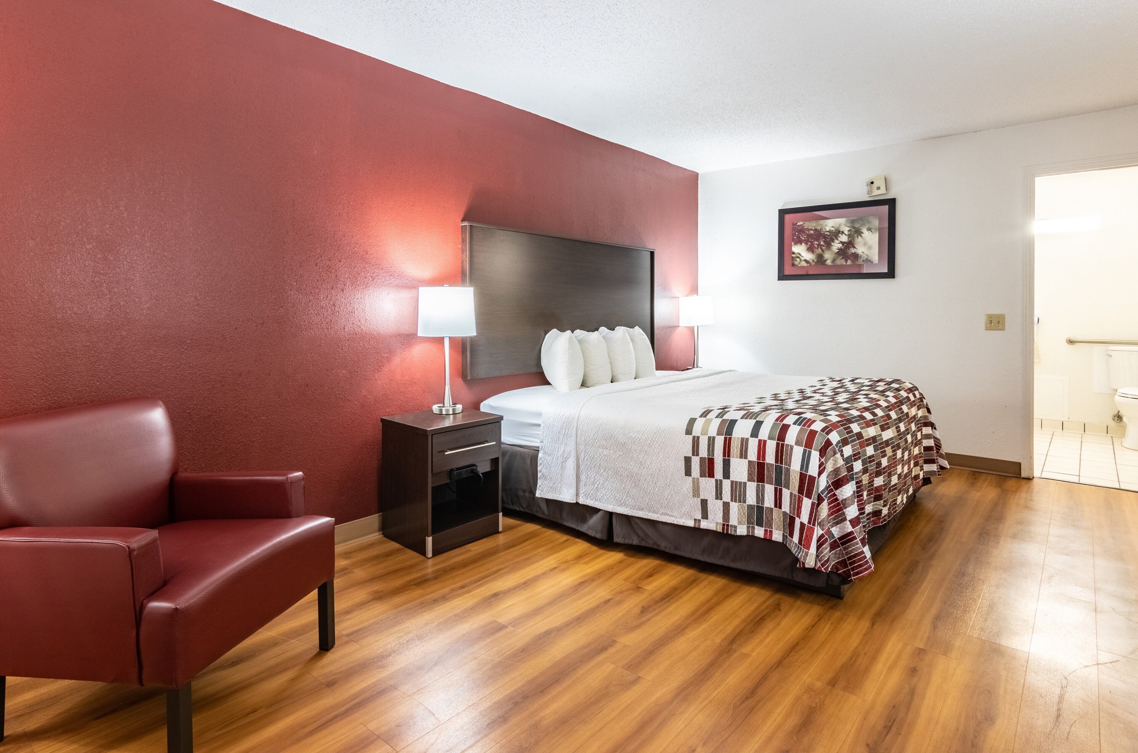 A hotel room with a bed, bedside table, lamp, chair, and a framed picture on the wall.