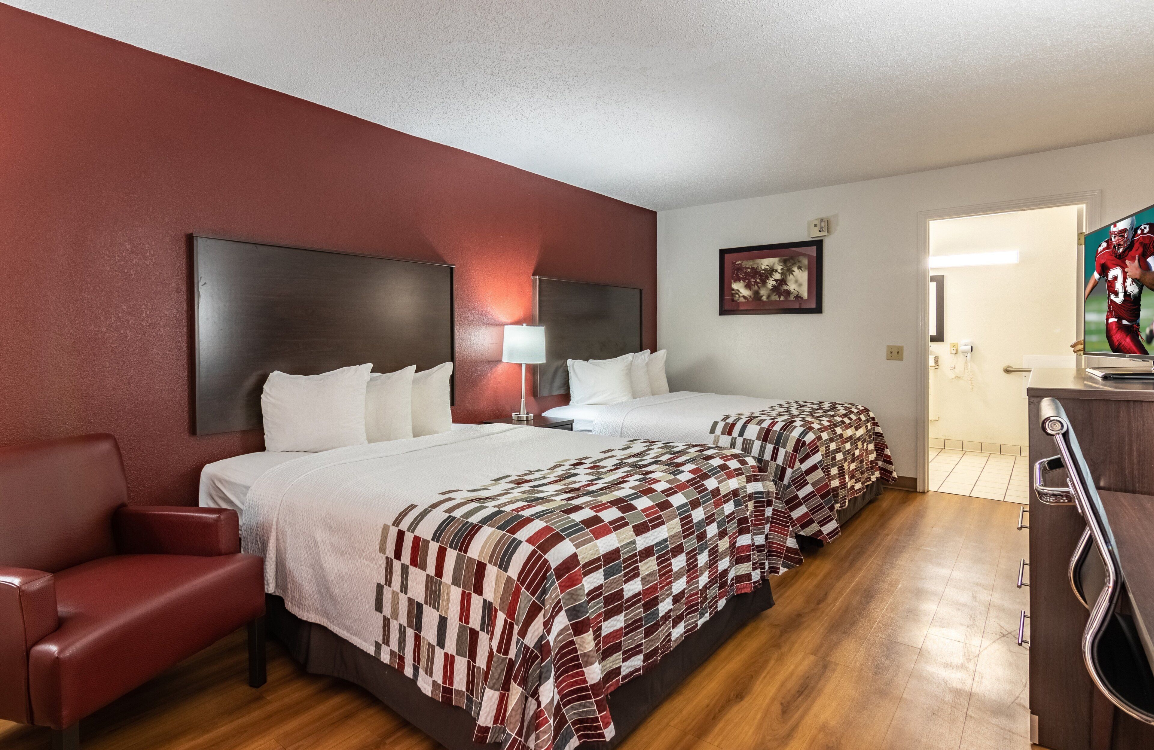 A hotel room with two beds, a red armchair, a desk with a mirror, and a TV.