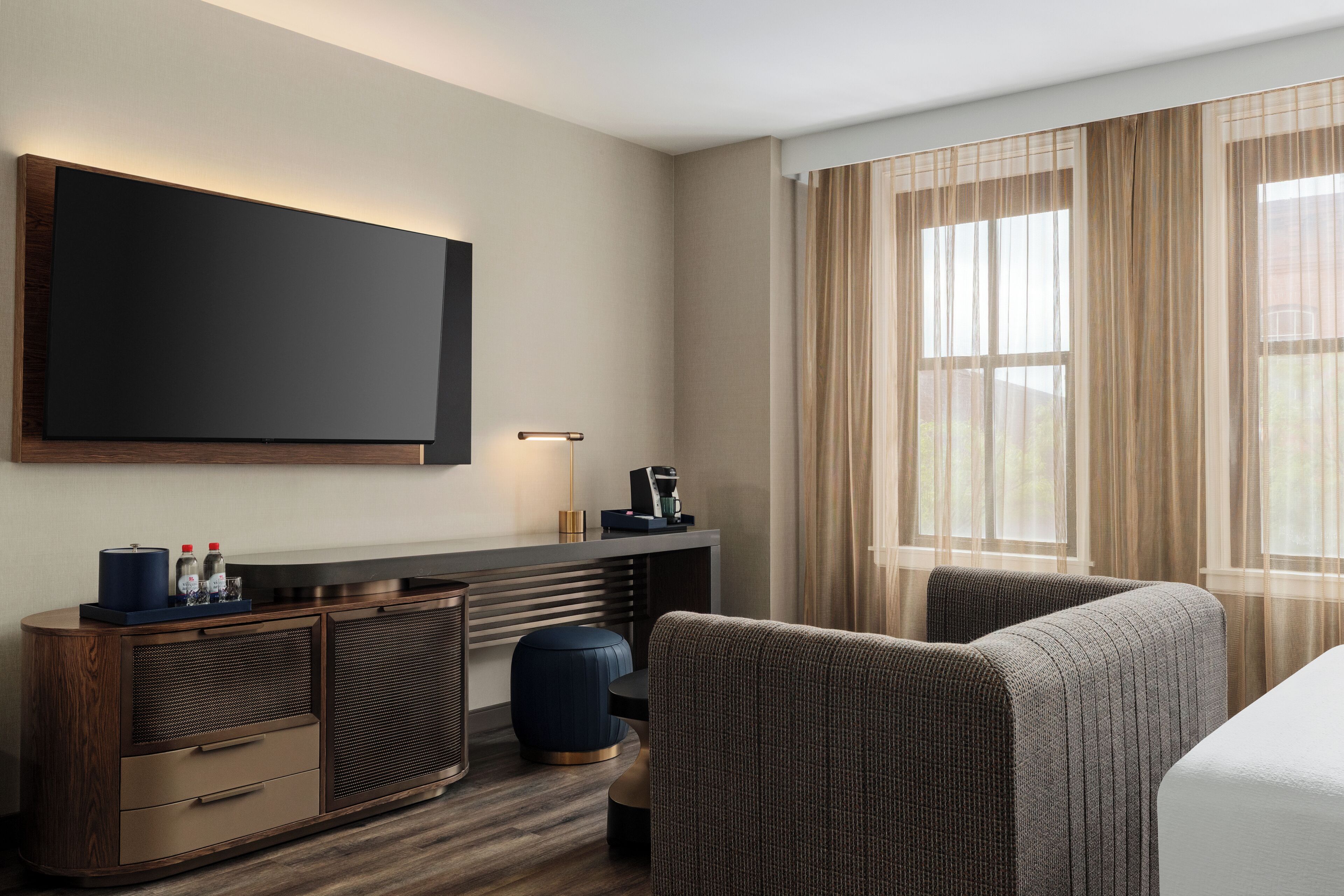A hotel room with a flat-screen TV mounted on the wall, a desk with a lamp, a coffee maker, and a seating area with a sofa and armchairs.