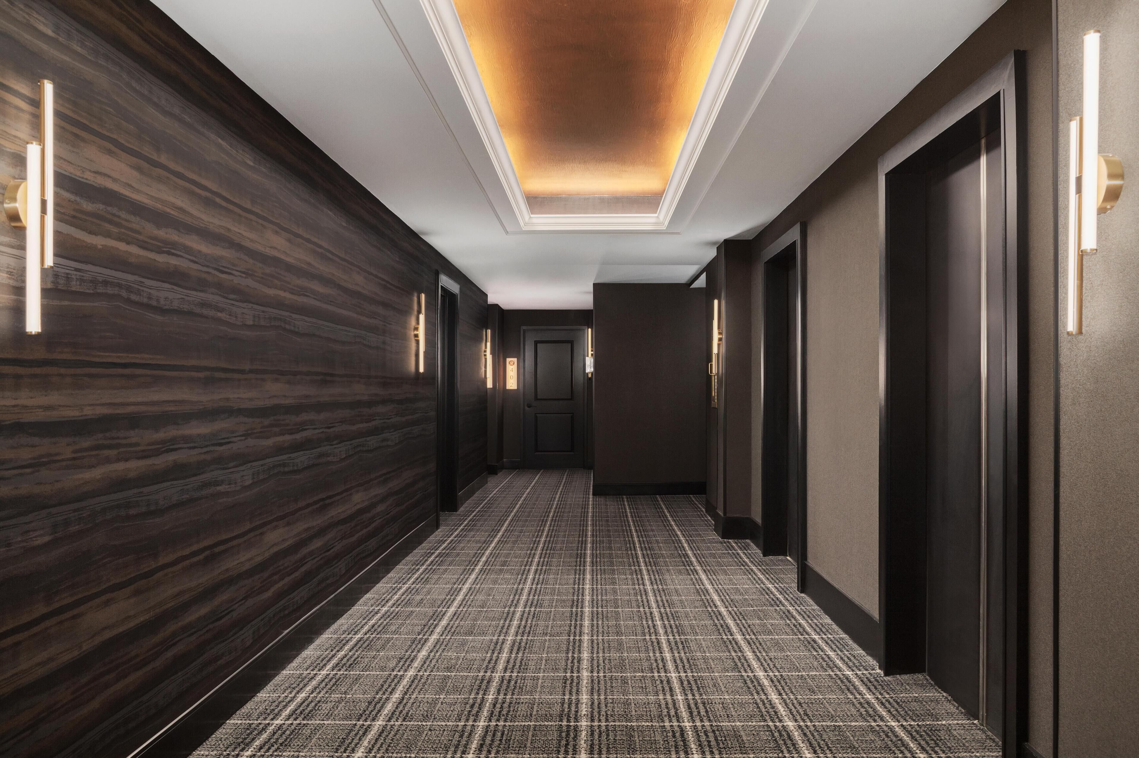 A modern hotel hallway with dark wood paneling, a checkered carpet, and a ceiling with recessed lighting.