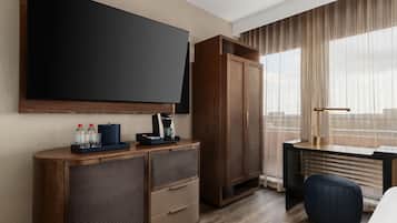 A hotel room with a large flat-screen TV, a minibar, a desk with a coffee maker, a wardrobe, and a view through sheer curtains.