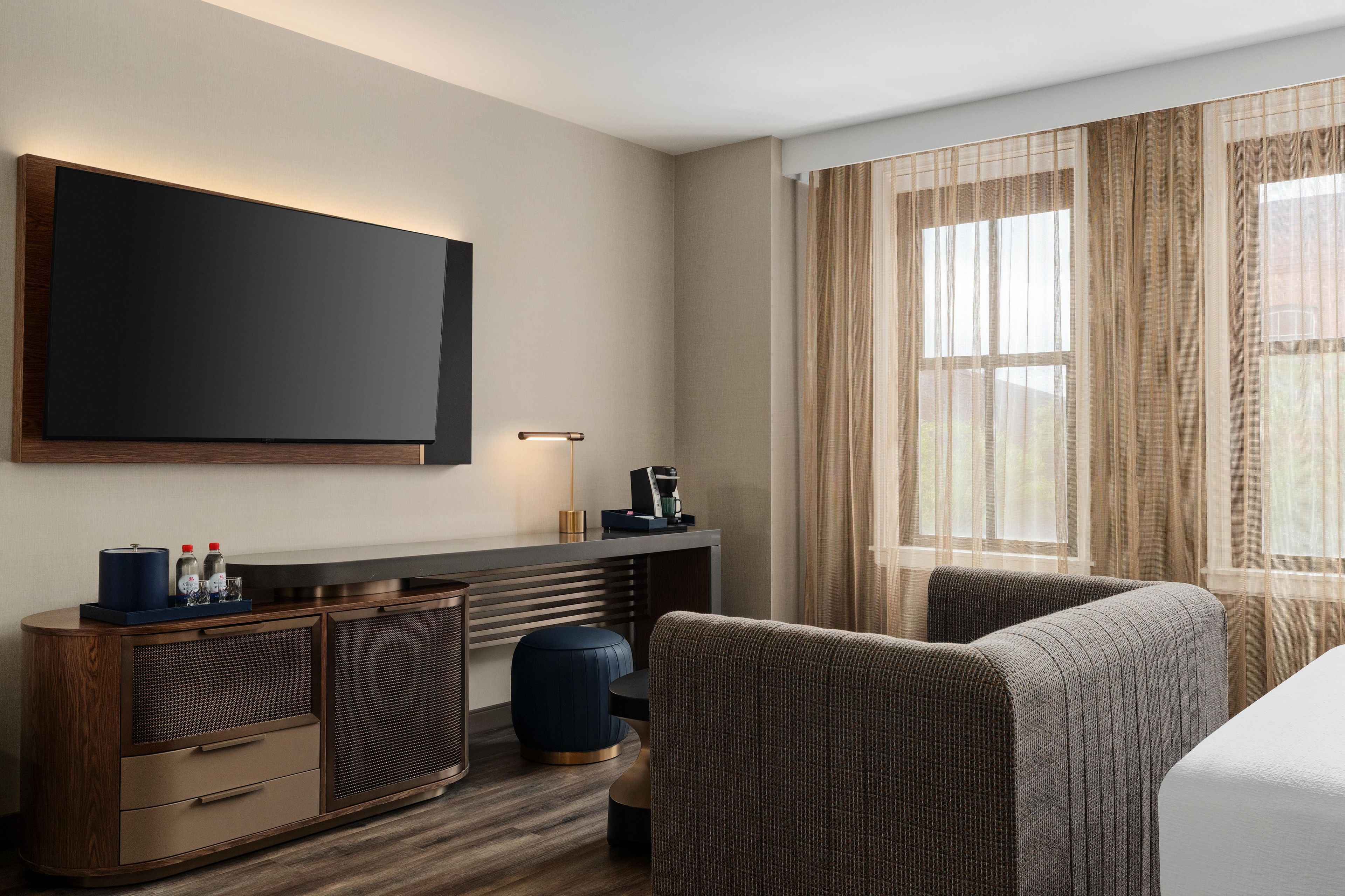 Habitación de hotel con televisor de pantalla plana, escritorio con lámpara, cafetera, sofá y ventana grande con cortinas translúcidas.