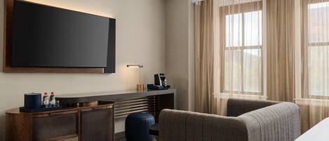 Habitación de hotel con televisor de pantalla plana, escritorio con lámpara, cafetera, sofá y ventana grande con cortinas translúcidas.