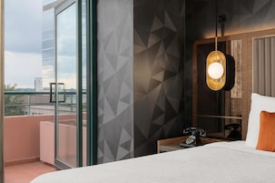 Habitación de hotel con un ventanal grande, cama con cabecera blanca, mesita de noche con teléfono y pared con diseño geométrico.