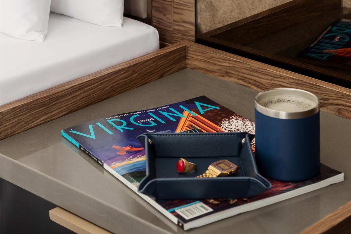 A neatly arranged hotel room with a bed, a bedside table with a book, a cup, and a small tray with a ring and a watch.