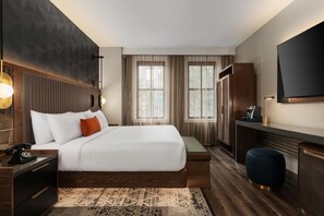 Una habitación de hotel moderna con una cama grande, mesitas de noche, un escritorio, una silla, un televisor y un ventanal amplio.