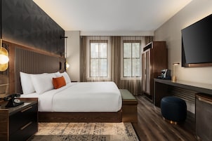Una habitación de hotel moderna con una cama grande, mesitas de noche, un escritorio, una silla, un televisor y un ventanal amplio.