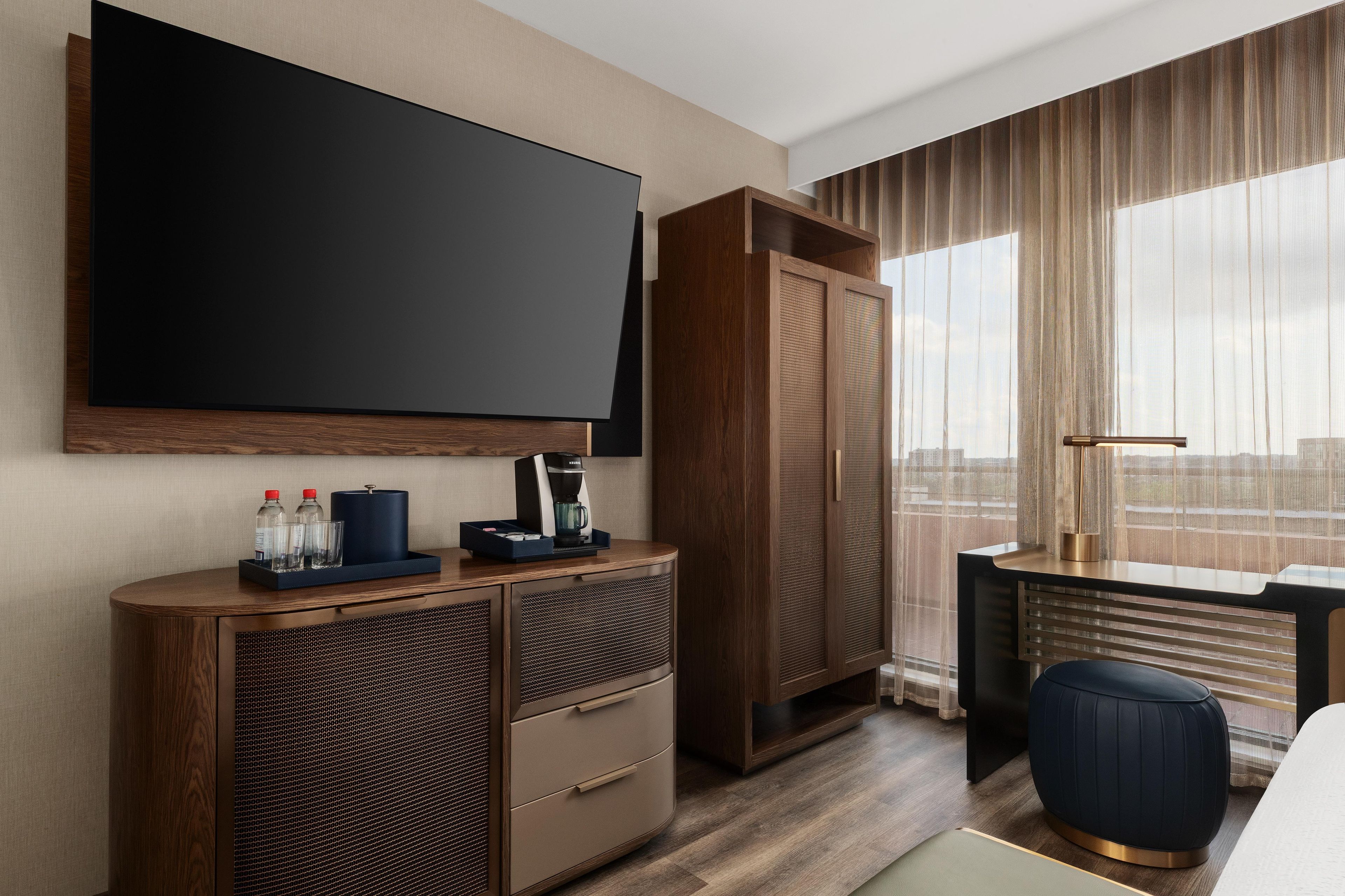 A hotel room with a large flat-screen TV, a minibar, a desk with a coffee maker, a wardrobe, and a view through sheer curtains.