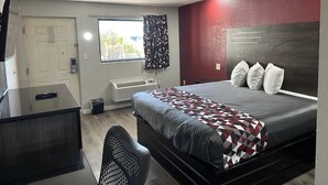 A bedroom with a bed, a desk, a chair, a window with curtains, and a radiator. - Rodeway Inn Jonesboro - Atlanta South (Forest Park)