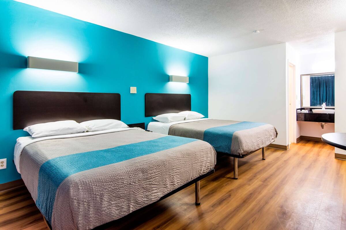 A hotel room with two beds, a wooden floor, and a bathroom with a sink and mirror.