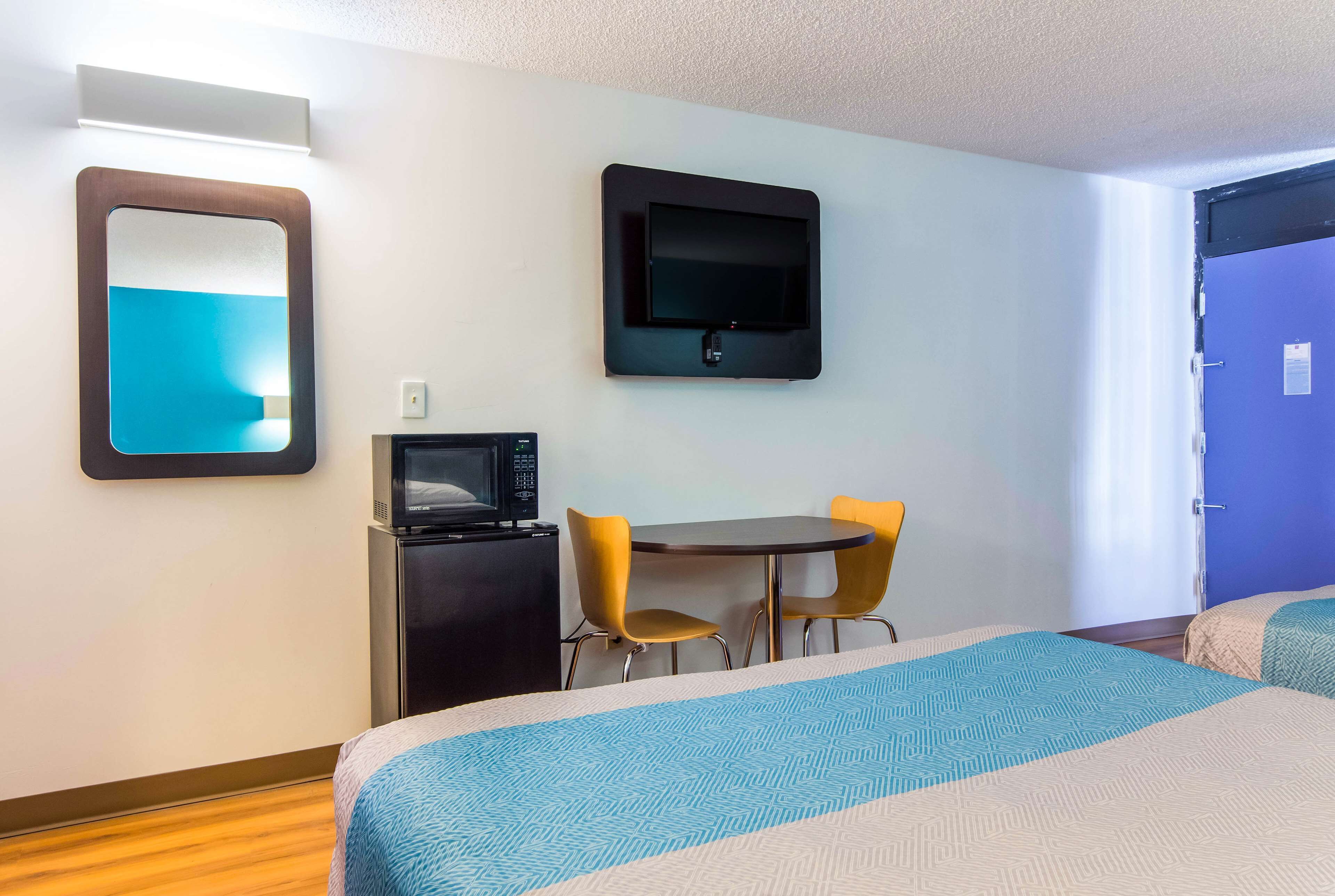 Een hotelkamer met een bed, een bureau met een magnetron, twee stoelen, een aan de muur bevestigde televisie en een deur met een blauwe omlijsting.