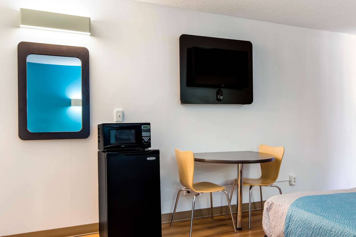 A compact hotel room with a bed, a small table with two chairs, a microwave, a flat-screen TV, and a mirror.
